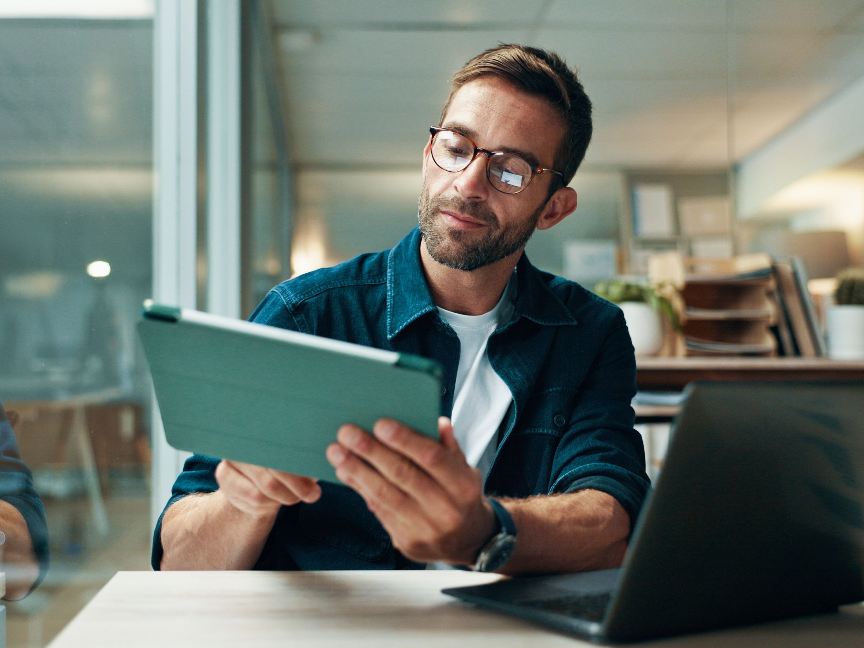 A professional man with glasses and beard wearing a navy button-up shirt sits at a desk, holding and looking at a tablet device in a modern office environment with glass walls and natural lighting.