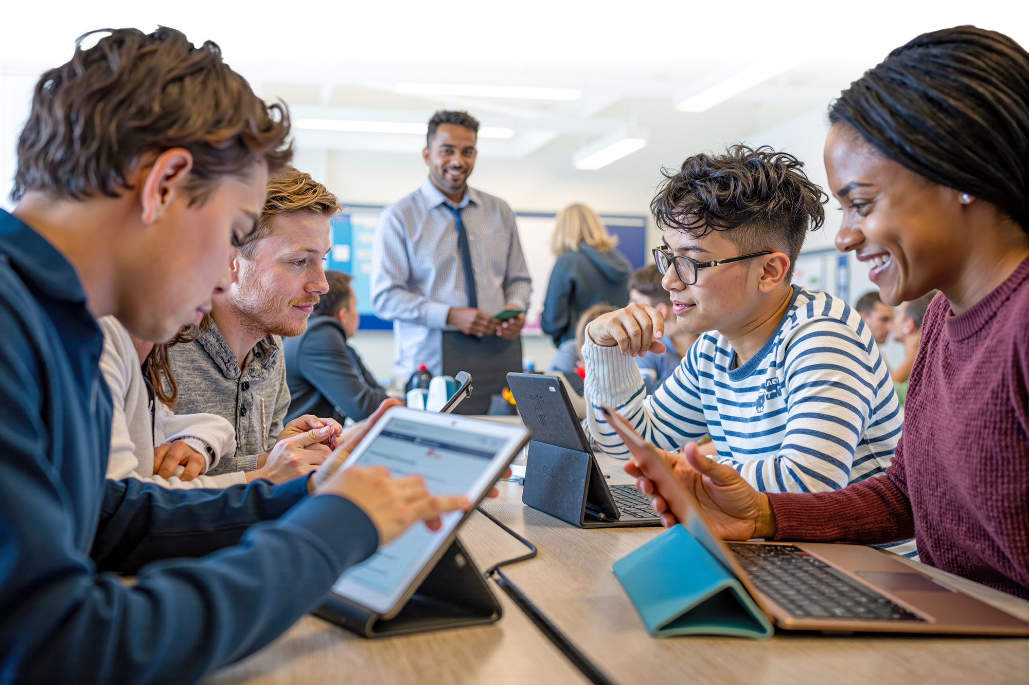 Students sitting around a table, working on tablets and smiling. In the distance a teacher watches with a smile.