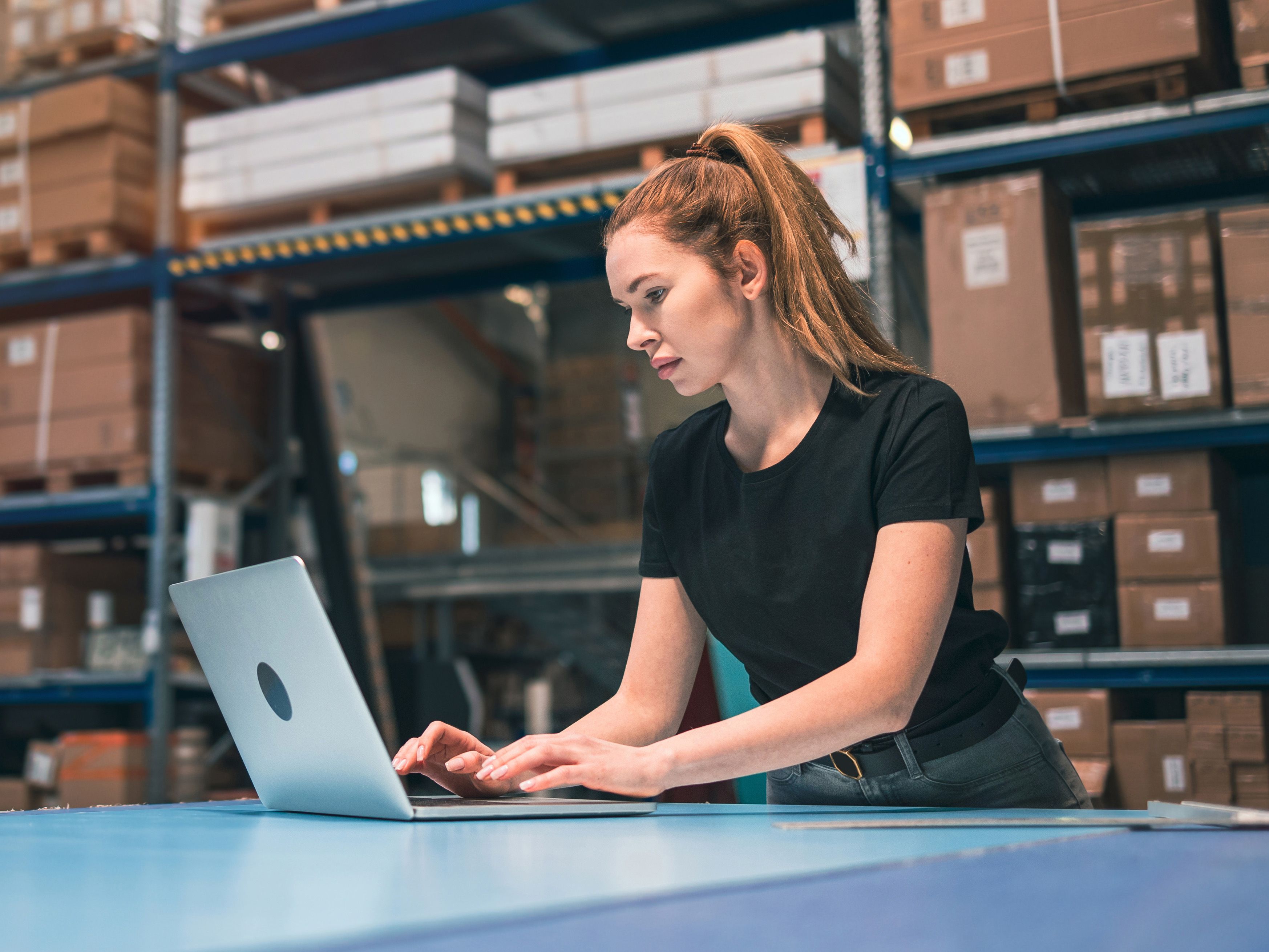 A woman standing at bench with a laptop, in front of shelves full of boxes.